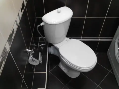 Установленный белый унитаз на тёмной плитке с подводкой воды сбоку
